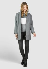 Grau melierter Wollmischmantel mit hohem Kragen, Druckknopfverschluss und seitlichen Taschen; getragen über einem gemusterten Pullover und schwarzen Slim-Fit-Jeans mit Stiefeletten.