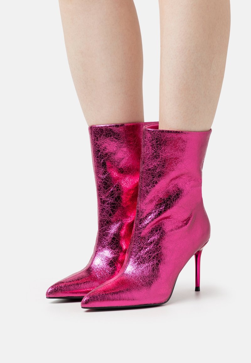 Bottes-ankle roses métalliques brillantes à bout pointu avec talons aiguilles portées sur des jambes nues contre un fond clair uni.