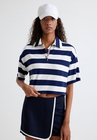 Marineblå og hvidstribet cropped polo skjorte med korte ærmer, featuring en knaplukket krave og en lille rød accent. Kombineret med en marineblå nederdel.