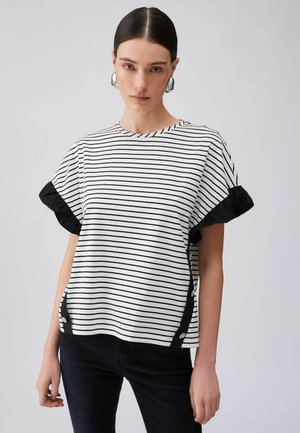 Giovane donna con capelli pettinati all'indietro che indossa orecchini a cerchio d'argento, una maglia a maniche corte a righe bianche e nere con dettagli neri e pantaloni neri.