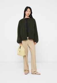 Woman wearing dark green fleece jacket, black top, beige wide-leg pants, pink flats, holding pale yellow handbag against white background.