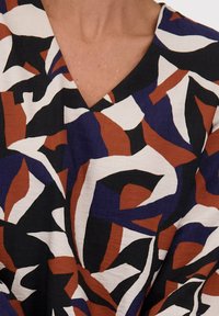 Close-up van een veelkleurig V-hals kledingstuk met abstracte zwarte, witte, roestkleurige en donkerblauwe vormen op textuurstof.