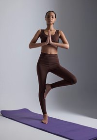 Woman in brown workout clothes balancing on one foot in tree yoga pose on purple mat against plain background.