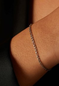 Zilveren kettingarmband om een pols, met langwerpige ovale schakels met een gladde textuur. Tegen een achtergrond van warme huid en donkere stof.