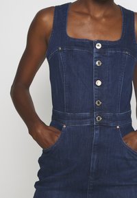 Tummansinisestä denimsarjasta valmistettu haalari, jossa on neliönmallinen kaula-aukko, nappikiinnitys edessä sekä kaksi sivutaskua. Teksturoitu kangas, jossa on sileä pinta.