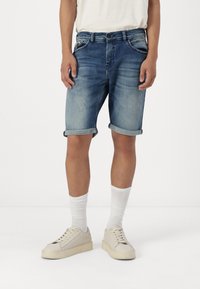 Shorts de mezclilla en azul claro con un acabado desgastado, dobladillos enrollados y diseño de cinco bolsillos. Combinados con calcetines blancos y zapatillas de color crema.