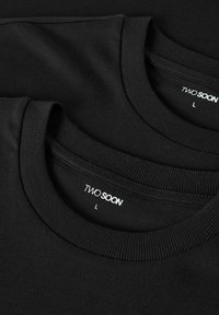 Černá bavlněná trička naskládaná na sobě, s kulatými výstřihy a žebrovanými manžetami. Vnitřní štítky nesou nápis "TWO SOON" a velikost "L". Měkká, hladká textura.