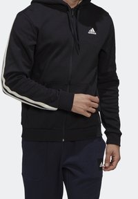 Homme portant un sweat à capuche zippé noir Adidas avec des bandes blanches sur les manches et un pantalon Adidas bleu marine, debout sur un fond clair.