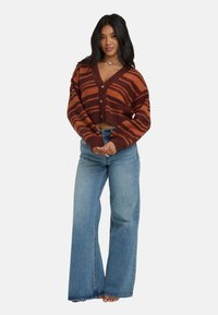 Braun und orange gestreifte cropped Strickjacke mit tiefem V-Ausschnitt und drei Knöpfen, kombiniert mit hellblauen weit geschnittenen Jeans. Weiche Strickstruktur.