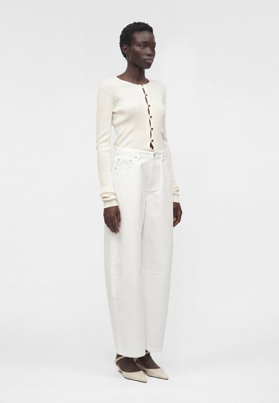 Γυναίκα που φοράει ένα εκρού ριμπ ζακέτα με ασύμμετρο κούμπωμα, φαρδιά λευκά παντελόνια με ψηλή μέση και μπεζ μυτερά τακούνια, στέκεται μπροστά από ένα λευκό φόντο.