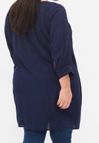 Mörkblå tunika med trekvartsärmar, subtil textur och sidoflikar. Tyget verkar lätt och något draperat.