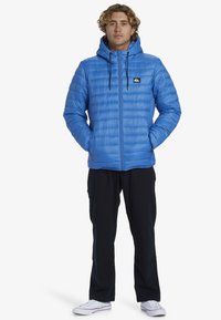 Blå pufferjakke med vandret quiltet design, hætte og lynlås foran. Har et logo patch på brystet; båret over sorte bukser og sneakers.