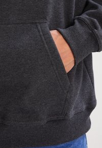 Ręka włożona do przedniej kieszeni ciemnoszarego hoodie z prążkowanymi mankietami, z widocznymi na dole niebieskimi dżinsami.