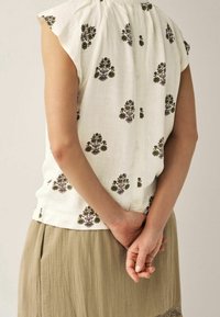 Biała bluzka z kwiatowym haftem w zieleni i brązie, z krótkimi rękawami, luźnego kroju. W połączeniu z długą, zwiewną, beżową spódnicą o fakturze.