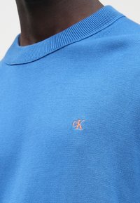 Približen posnetek osebe, ki nosi modro pleteno majico z majhnim vezenim logotipom "cK" v oranžni barvi na prsih.