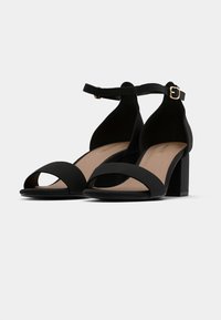 Mustat korotetut sandaalit, jotka on valmistettu pehmeästä kankaasta. Niissä on nilkkahihna soljella, avonainen kärki ja tukevat paksut korot.