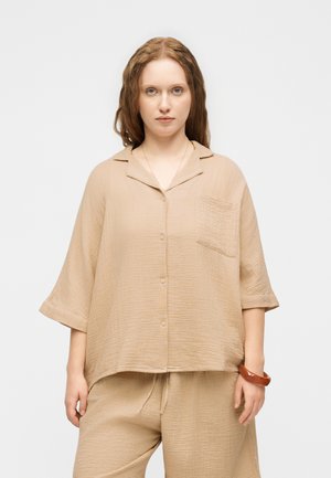 Frau mit heller Haut und langen welligen Haaren, die ein lockeres beiges Hemd mit Knöpfen und passende Hosen trägt, steht vor einem schlichten Hintergrund.