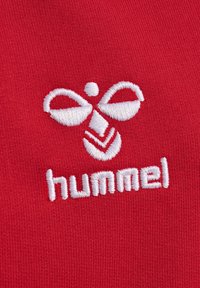Tessuto rosso con un logo bianco ricamato, caratterizzato da un design simmetrico e la parola "hummel" in un font moderno.