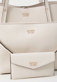 Trois sacs en cuir beige avec texture de galet, arborant le logo doré "GUESS". Comprend un grand tote et un petit portefeuille avec fermeture à rabat et détails de couture.