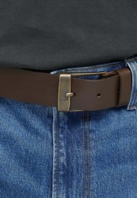 Brauner Ledergürtel mit einer rechteckigen Messingschnalle, getragen über blauen Jeans mit orangefarbenen Nähten. Nahaufnahme des unteren Oberkörpers.