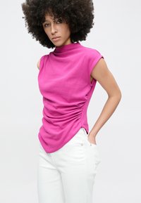 Fuchsia mouwloze top met een hoge hals, voorzien van zijsamenplooingen en een zachte textuur, gecombineerd met witte nauwsluitende broek.