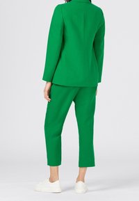 Zelený upravený blejzr a odpovídající zkrácené kalhoty vyrobené z hladkého materiálu. Obsahuje límec, strukturovaný střih a jemné stehové detaily.