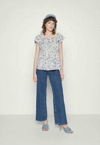 Top com padrão de flores e mangas em babados, cintura peplum, combinado com jeans Azul de perna larga e saltos azuis a condizer. Fundo neutro.