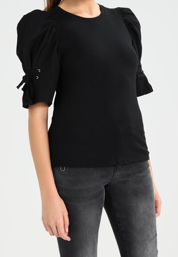 Donna che indossa una maglietta nera a maniche corte con maniche a sbuffo e dettagli con lacci, abbinata a jeans neri scoloriti su uno sfondo semplice.