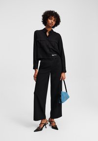 Camicia nera a maniche lunghe con due tasche sul petto, abbinata a pantaloni neri a gamba larga. Tiene in mano una borsa blu e indossa décolleté nere a punta.