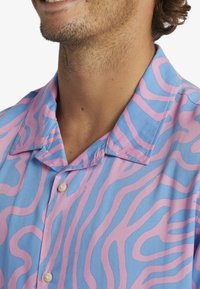 Man in een blauw en roze gestreepte overhemd met korte kraag, die een deel van zijn nek en glimlach laat zien.