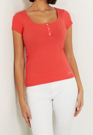 Mujer con una camiseta ajustada coral de manga corta con tres botones decorativos y pantalones blancos sobre un fondo liso.