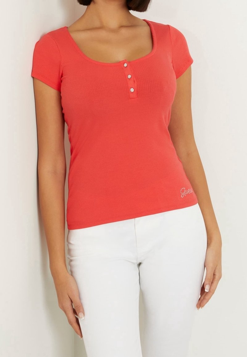 Femme portant un haut corail moulant à manches courtes avec trois boutons décoratifs et un pantalon blanc, sur un fond uni.