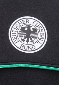 Στρογγυλό ασπρόμαυρο λογότυπο του Deutscher Fussball Bund με έναν αετό στο κέντρο σε μαύρο ύφασμα πάνω από μια πράσινη λωρίδα.