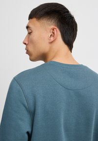Maglione teal con scollatura rotonda, polsini a costine e una texture liscia. Presenta un particolare dettaglio di cucitura sul retro.