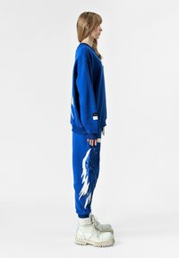 Sudadera azul y pantalones de chándal a juego con un patrón de alas en blanco, fabricados en un tejido suave. Unos botines blancos gruesos completan el conjunto.