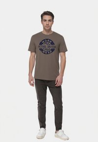 Oliwkowy bawełniany T-shirt z okrągłym granatowym napisem "WORK PETROL DRIVEN WEAR." W zestawie z ciemnoszarymi dżinsami i białymi sneakersami.