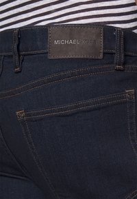 Zbliżenie na ciemnoniebieskie dżinsy z widocznymi kieszeniami i metką Michael Kors na pasku, zestawione z pasiastą koszulą powyżej.