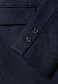 Tmavomodrý vlnený materiál zobrazuje detail rukávu saka so dvěma černými čtyřdírovými knoflíky a strukturovaným povrchem.