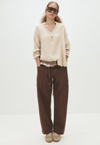 Beige Struktur-Oberteil mit V-Ausschnitt und ausgefranstem Halsausschnitt, kombiniert mit braunen, locker sitzenden Hosen mit Seitentaschen und einem geflochtenen Gürtel.