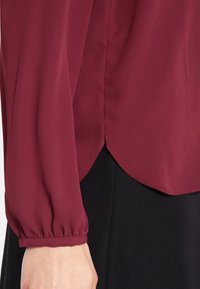 Blouse bordeaux à manches longues avec manches bouffantes, poignets plissés et ourlet arrondi. Le tissu semble lisse et léger. Portée sur un vêtement noir.