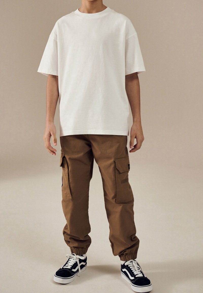 Personne portant un t-shirt blanc uni et ample, un pantalon cargo marron avec des poches latérales et des baskets noires et blanches, debout sur un fond beige.