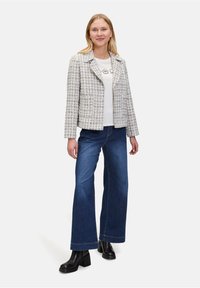 Geweven geruite jas in lichtgrijs en crème, gedragen over een wit shirt, gecombineerd met blauwe flared jeans en zwarte hielenlaarzen.