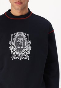 Temno moderna dukserica z rdečim šivanjem, z našitim belim logotipom, ki vključuje dekorativni ščit ter besedilo "HILFIGER" na dnu.