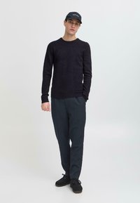 Pulover negru din tricot, pantaloni gri închis și pantofi negri. Modelul poartă o șapcă neagră cu text alb. Design simplu, textură moale.