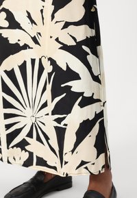 Maxi-jurk met bloemenpatroon in zwart en crèmekleurige stof, met zijsplitsen. Gecombineerd met zwarte instappers voor een harmonieuze uitstraling.