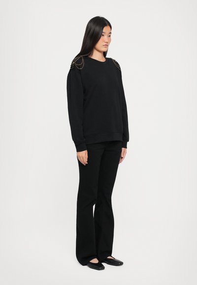 Czarna bluza z okrągłym dekoltem, długimi rękawami i metalicznymi łańcuszkami na ramionach, zestawiona z czarnymi spodniami w dzwony i balerinami.