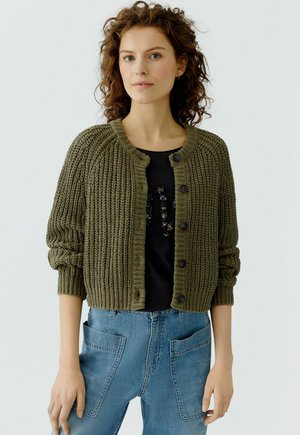 Frau trägt eine grob gestrickte olivgrüne Knopfstrickjacke über einem schwarzen Oberteil und eine hoch taillierte blaue Jeans mit großen vorderen Taschen.