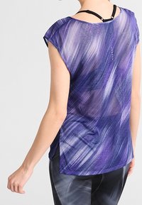 Vrouw draagt een doorschijnende paarse, patroonloze, mouwloze top over een zwarte beha en donker patroonleggings, met de rug naar voren en de linkerarm gebogen.