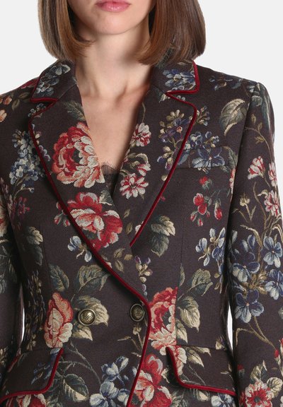 Gros plan d'un blazer à motif floral foncé avec passepoil rouge et boutons en laiton porté par une femme aux cheveux bruns mi-longs.