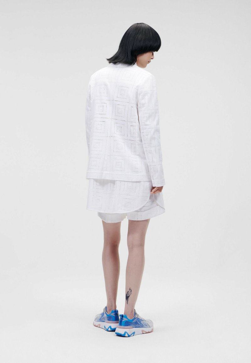 Weißes Blazer- und Rock-Set aus strukturiertem Stoff mit geometrischem Quadratmuster. Kombiniert mit mehrfarbigen Sneakers.